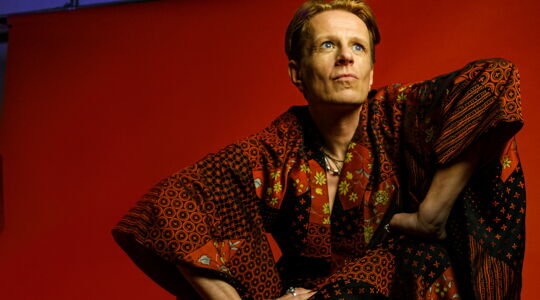 Portrait von Alexander Scheer vor rotem Hintergrund. Er hat blaue Augen, kurze blonde Haare und trägt einen rot-schwarz gemusterten Kimono.